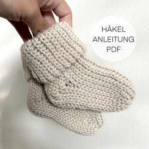 Op de afbeelding: Een paar beige gehaakte babyschoentjes met een gevouwen boord. De schoentjes worden in een hand gehouden. De tekst "HÄKEL ANLEITUNG PDF" is zichtbaar in de afbeelding.