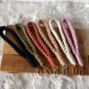 Puede incluir: Una colección de pulseras de macramé hechas a mano en varios colores, incluyendo negro, verde oliva, terracota, beige, crema y rosa. Cada pulsera presenta un diseño trenzado y un cierre plateado o dorado.