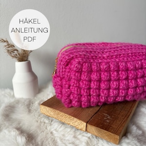 May include: A bright pink crocheted zippered pouch with a gold zipper. The pouch is sitting on a wooden surface.