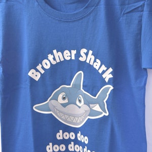 Puede incluir: Una camiseta azul con un tibur&oacute;n de dibujos animados blanco y el texto "Brother Shark doo doo doo doo doo" impreso en ella.
