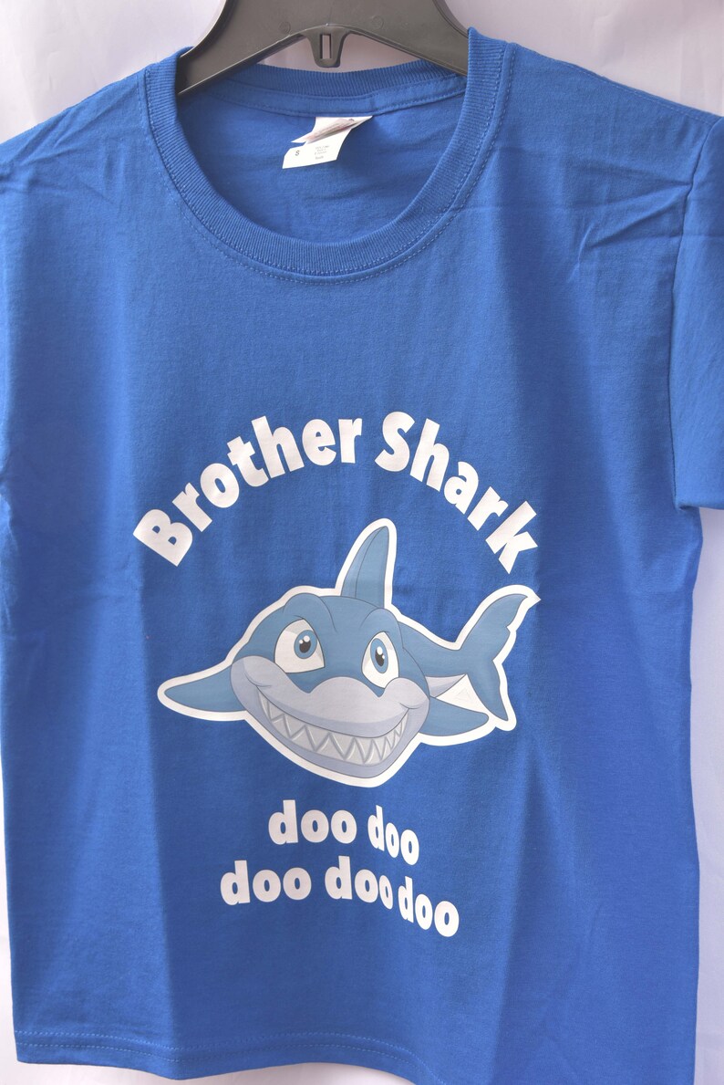 Puede incluir: Una camiseta azul con un tibur&oacute;n de dibujos animados blanco y el texto "Brother Shark doo doo doo doo doo" impreso en ella.