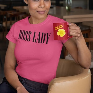 Może przedstawiać: R&oacute;żowy T-shirt z nadrukiem "BOSS LADY" w kolorze czarnym. Osoba na zdjęciu trzyma czerwony kubek z napisem "SUCK IT UP BUTTERCUP" i ż&oacute;łtym wzorem kwiatu.
