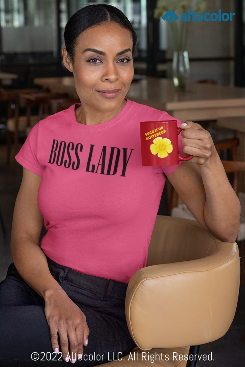 Może przedstawiać: R&oacute;żowy T-shirt z nadrukiem "BOSS LADY" w kolorze czarnym. Osoba na zdjęciu trzyma czerwony kubek z napisem "SUCK IT UP BUTTERCUP" i ż&oacute;łtym wzorem kwiatu.