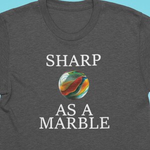 Puede incluir: Una camiseta gris oscuro con texto blanco que dice "SHARP AS A MARBLE". Una canica colorida está centrada entre las dos líneas de texto.
