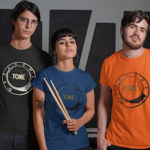 Puede incluir: Tres personas con camisetas con un gráfico de un mando de tono. Las camisetas son negras, azules y naranjas. La persona del medio sostiene dos baquetas.