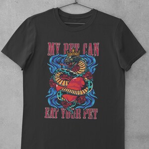 Puede incluir: Camiseta negra con un gráfico colorido de una serpiente coronada enrollada alrededor de un corazón rojo. El texto "MY PET CAN EAT YOUR PET" se muestra arriba y abajo del gráfico en una fuente vintage.