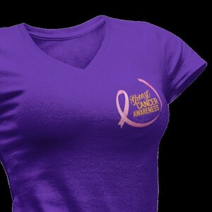 Puede incluir: Camiseta morada de cuello en V con una cinta rosa y el texto "Breast Cancer Awareness" en letras doradas.
