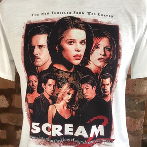 Può includere: Maglietta bianca con una grafica rossa e nera del film Scream 2. La grafica presenta i personaggi principali del film e il testo "THE NEW THRILLER FROM WES CRAVEN" e "SCREAM 2". Il testo sotto la grafica recita "Someone has taken their love of sequels one step too far."