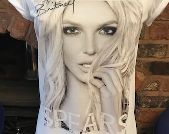 May include: White t-shirt with a black and white photo of Britney Spears on the front. The photo shows Britney Spears looking at the camera with her hair covering part of her face. The text "Britney Spears" is printed on the shirt in black.