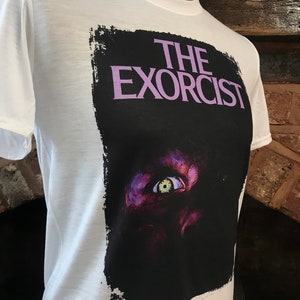 May include: White t-shirt with a black and white graphic print of the movie "The Exorcist". The graphic features a close-up of a demonic eye with a yellow iris.