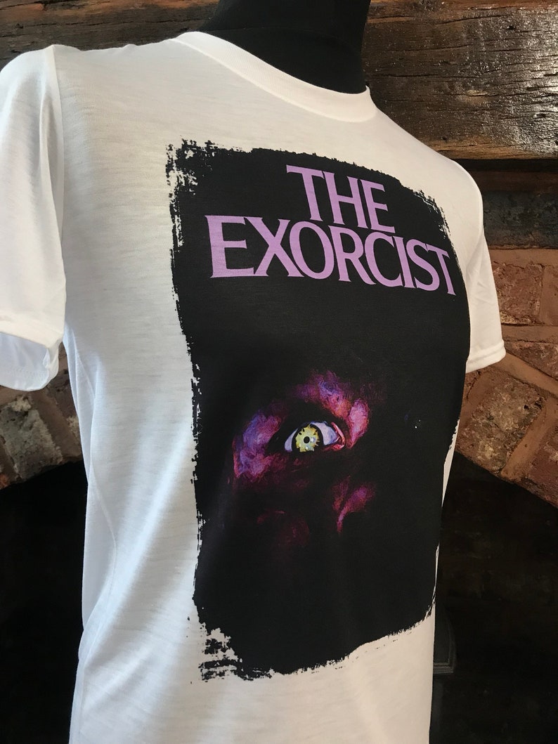 May include: White t-shirt with a black and white graphic print of the movie "The Exorcist". The graphic features a close-up of a demonic eye with a yellow iris.
