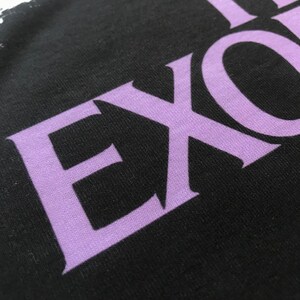 May include: Close-up of a black t-shirt with the word "THE" and the word "EXO" in a purple font.