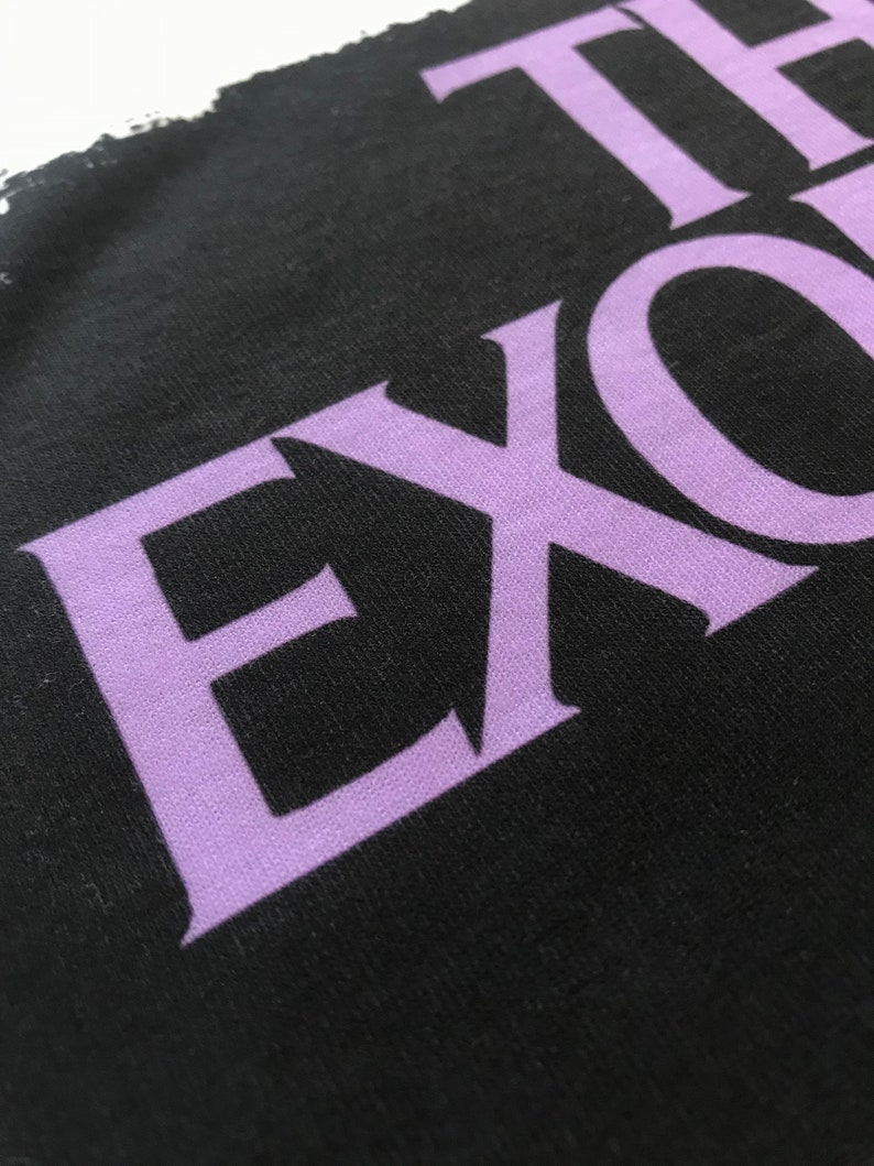 May include: Close-up of a black t-shirt with the word "THE" and the word "EXO" in a purple font.