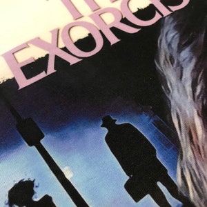 May include: A vintage-style graphic tee with a dark blue background featuring the text "THE EXORCIST" in a light purple font. The image shows two figures walking away from the viewer in silhouette, with a streetlight and a moon in the background.