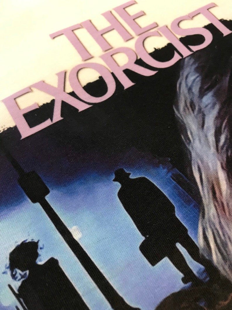 May include: A vintage-style graphic tee with a dark blue background featuring the text "THE EXORCIST" in a light purple font. The image shows two figures walking away from the viewer in silhouette, with a streetlight and a moon in the background.