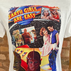 Puede incluir: Camiseta blanca con un estampado gráfico de un collage de imágenes de la película "Earth Girls Are Easy". Las imágenes incluyen los personajes principales, una nave espacial y el título de la película.