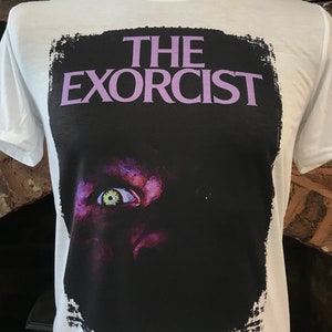 May include: White t-shirt with a black and white image of a demonic eye and the text "THE EXORCIST" in purple.