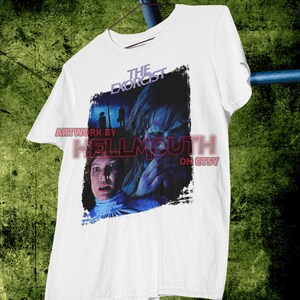May include: White t-shirt with a graphic print of a scene from the movie "The Exorcist". The graphic features a woman with a scared expression and a young girl with a demonic look in her eyes. The text "The Exorcist" is printed in black above the graphic.