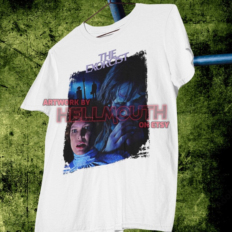 May include: White t-shirt with a graphic print of a scene from the movie "The Exorcist". The graphic features a woman with a scared expression and a young girl with a demonic look in her eyes. The text "The Exorcist" is printed in black above the graphic.