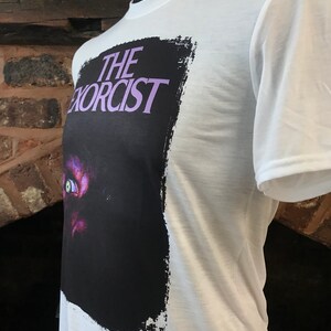 May include: White t-shirt with a black and white graphic print of the movie "The Exorcist". The graphic features a close-up of a demonic eye with a purple hue.