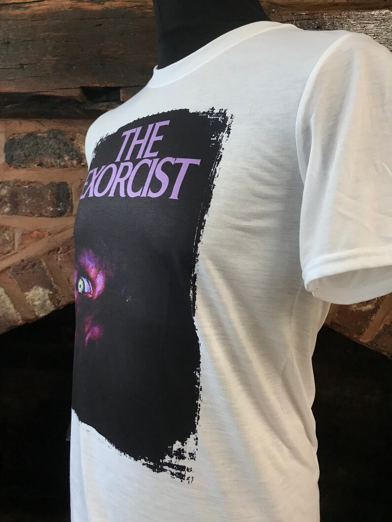 May include: White t-shirt with a black and white graphic print of the movie "The Exorcist". The graphic features a close-up of a demonic eye with a purple hue.