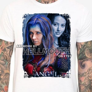 Peut inclure: Un t-shirt blanc avec un imprimé graphique de deux femmes aux cheveux bleus et rouges. Le texte "Artwork by Hellmouth on Etsy" et "Angel" sont imprimés sur le t-shirt.