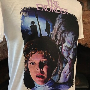 May include: White t-shirt with a graphic print of a scene from the movie "The Exorcist". The graphic features a close-up of the possessed girl and the demon, with a shadowy figure in the background.