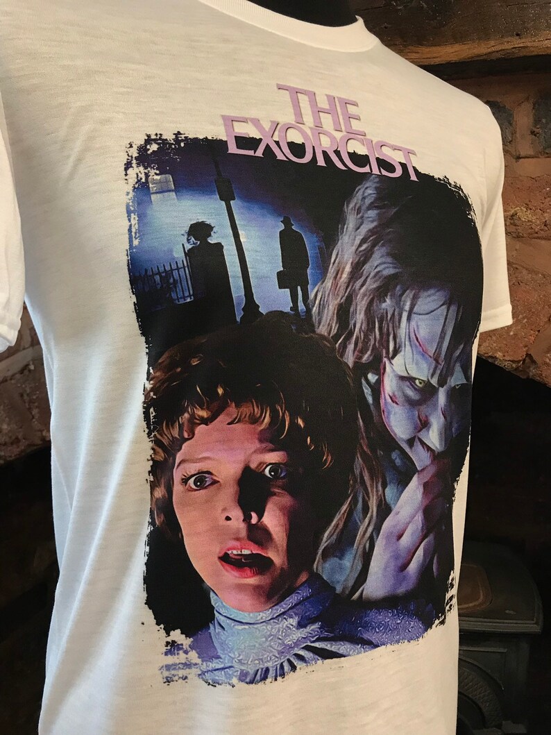 May include: White t-shirt with a graphic print of a scene from the movie "The Exorcist". The graphic features a close-up of the possessed girl and the demon, with a shadowy figure in the background.