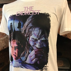 May include: White t-shirt with a black and white graphic print of a scene from the movie "The Exorcist". The graphic features a young girl with a scared expression and a demon with a menacing look.