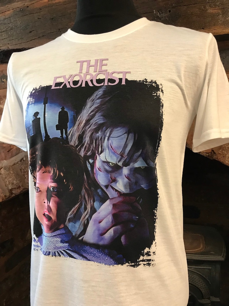 May include: White t-shirt with a black and white graphic print of a scene from the movie "The Exorcist". The graphic features a young girl with a scared expression and a demon with a menacing look.