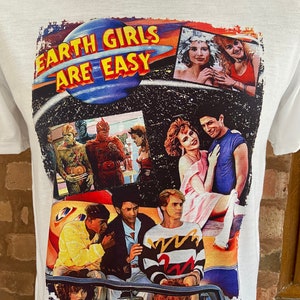 Puede incluir: Camiseta blanca con un estampado de collage colorido con el texto "Earth Girls Are Easy" e imágenes de la película del mismo nombre. La impresión incluye escenas de los personajes principales, incluyendo una nave espacial, un automóvil y un grupo de extraterrestres.