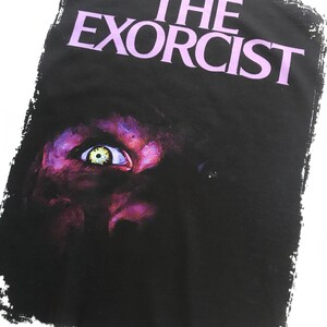 May include: A black and white graphic t-shirt with the text "THE EXORCIST" in purple lettering. The image features a close-up of a demonic eye.