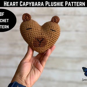 May include: A heart-shaped, brown crochet plushie with a capybara face, including small ears and closed eyes. The text "HEART CAPYBARA PLUSHIE PATTERN" is at the top, and "PDF CROCHET PATTERN" is in a black circle. A butterfly logo is in the bottom right.