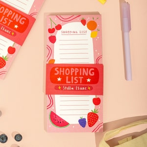 May include: Two pink shopping list pads with a fruit theme. The top pad has the text "SHOPPING LIST" with a star on either side and the name "Isaac" written below. The bottom pad has the text "SHOPPING LIST" with a star on either side and the name "Stella Isaac" written below. Both pads feature a variety of fruits drawn on them, including strawberries, cherries, lemons, and watermelon.