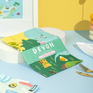 May include: A green tea towel with a map of Devon, England. The tea towel features the text "DEVON Teatowel Stella Isaac" and includes the names of towns and cities in Devon, including Exeter, Crediton, Tiverton, Topsham, and Newton Abbot.