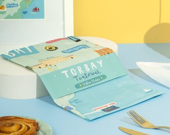 Può includere: Un asciugamano da cucina blu con una mappa di Torbay, in Inghilterra. L'asciugamano da cucina presenta il testo "Torbay Tea Towel" e "Stella Isaac".