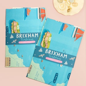 May include: Two blue tea towels with a map of Brixham, England. The towels feature a blue and white design with red accents. The text "Brixham Teatowel Stella Isaac" is printed on the towels.