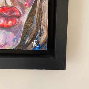 May include: A close-up of a framed painting of a colorful abstract portrait. The painting is signed with the letter "E" in the bottom right corner.
