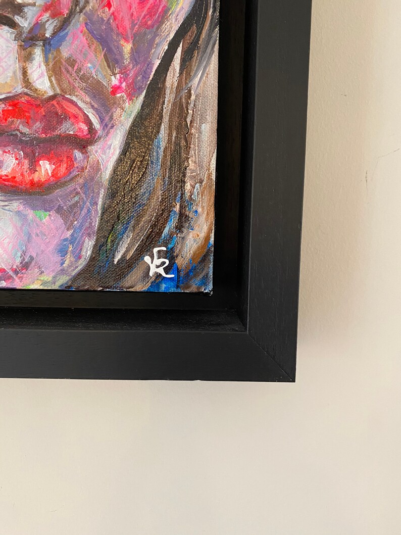 May include: A close-up of a framed painting of a colorful abstract portrait. The painting is signed with the letter "E" in the bottom right corner.