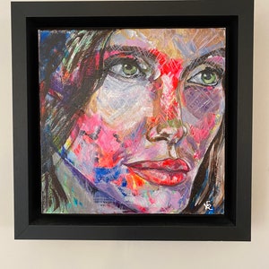 May include: A framed painting of a woman's face with green eyes and red lips. The painting is done in a colorful abstract style with splashes of pink, blue, and green.