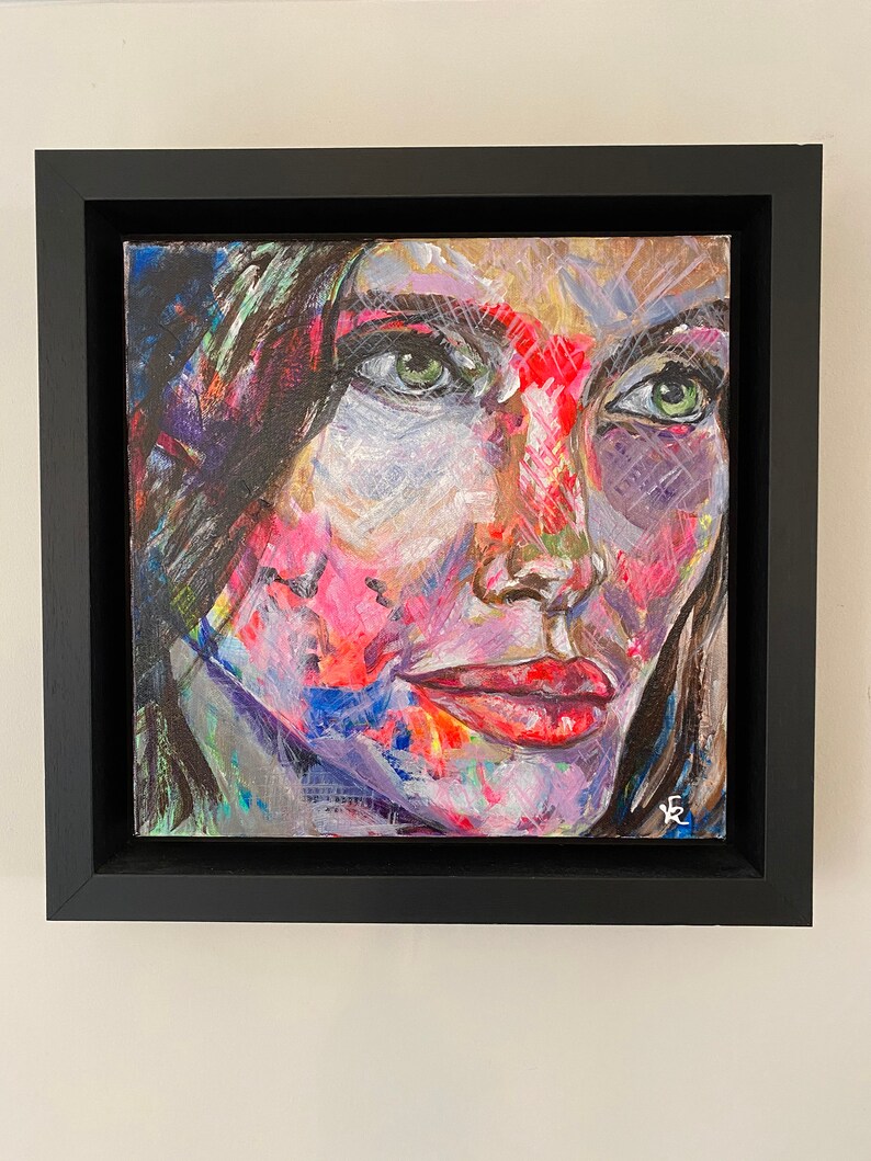 May include: A framed painting of a woman's face with green eyes and red lips. The painting is done in a colorful abstract style with splashes of pink, blue, and green.