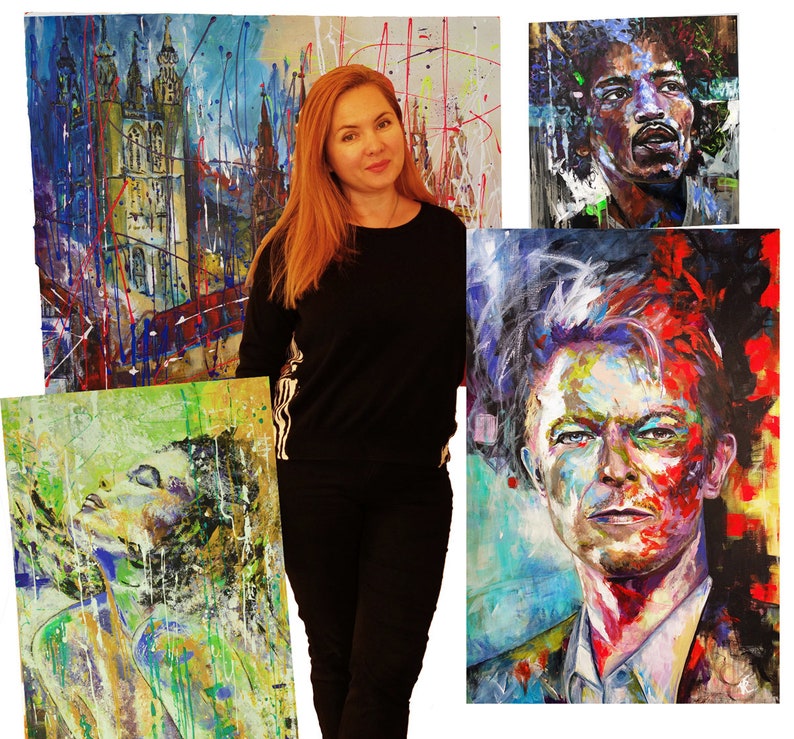 May include: A woman stands in front of four colorful paintings. The paintings are abstract and feature a variety of colors and textures. The woman is wearing a black shirt and jeans.