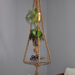 Può includere: Un portapiante in macramè in corda beige, con un ripiano in vetro trasparente e due piante in vaso con foglie verdi. Un grappolo di uva viola poggia sul ripiano. Appeso contro un muro grigio.