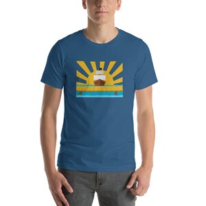 Op de afbeelding: Een blauw t-shirt met een afbeelding van een schip dat naar een gele zon met stralen vaart, over een houten dok en blauw water.