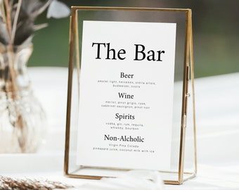 Puede incluir: Un cartel con marco dorado, fondo blanco y texto negro que enumera el menú del bar. El menú incluye cerveza, vino, licores y opciones sin alcohol. El cartel está sobre una mesa con mantel blanco.