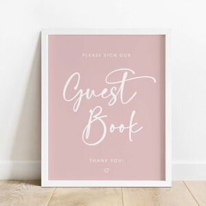 May include: A white framed sign with a pink background. The sign says "Please sign our Guest Book. Thank you!" in white cursive text.