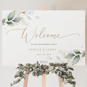 May include: A white welcome sign with green and gold foliage and the text "Welcome to our engagement party Ashley & James July 24 2024 Wasing Barn Denver, Col."
