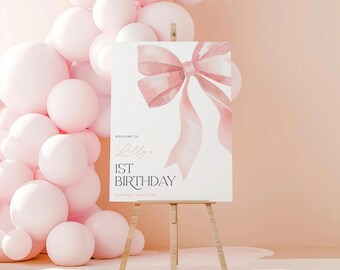 May include: A white sign on a wooden easel with a watercolor pink bow illustration. The sign reads "Welcome to Lilly's 1st Birthday" with the date April 26th. Pink balloons are in the background.