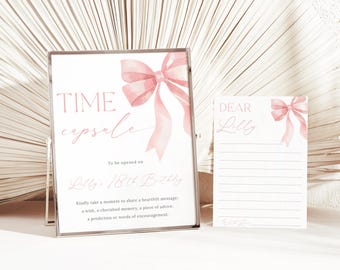 May include: A pink and white time capsule sign with a watercolor bow and the text "TIME capsule" and "To be opened on Lilly 18th Birthday". A white card with the text "DEAR Lilly" is next to the sign.