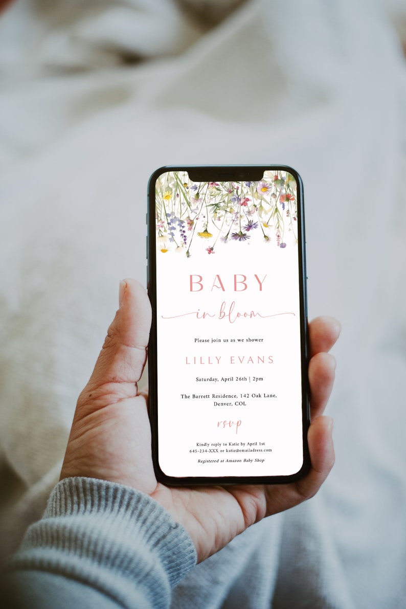 May include: A baby shower invitation on a smartphone screen with a floral design. The invitation reads "Baby in Bloom" and includes the name "Lilly Evans", the date and time of the shower, the location, and RSVP information.